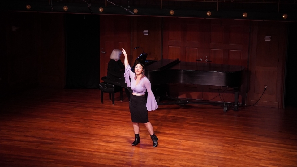 a young woman sings on stage in a purple blouse and black skirt