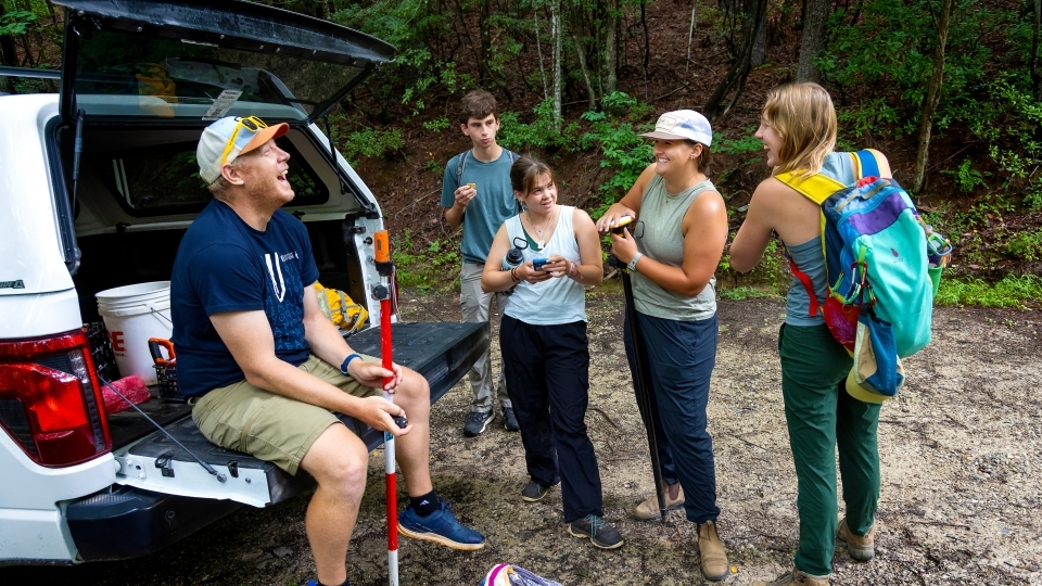 a group of students with their professor gathered around a truck in the woods wearing hiking gear