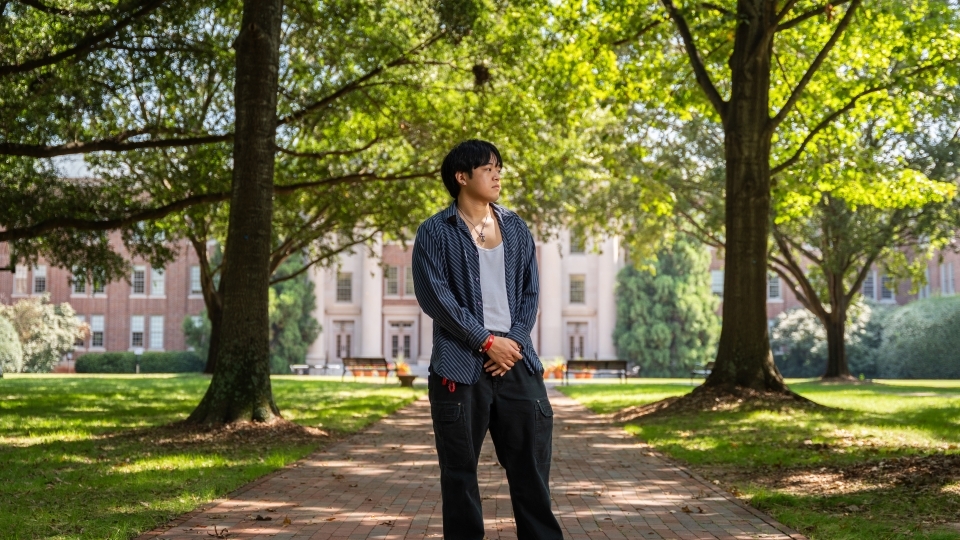 a young man stands on a college campus looking to his side in front of a large building surrounded by trees