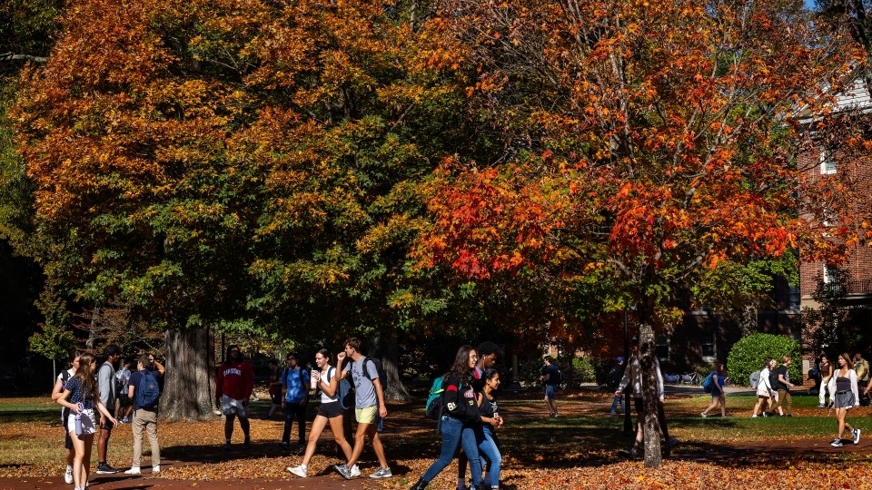 Students wearing backpacks walk past bright autumn foliage on the chambers lawn