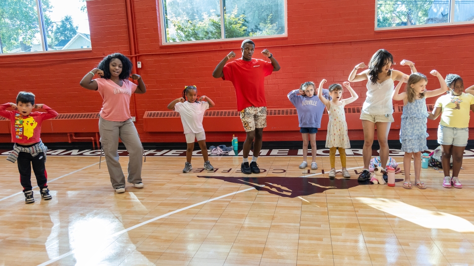 A group of diverse children and young adult leaders stand in a row on a gymnasium floor, smiling and flexing their muscles in a strong pose against a red and white wall.