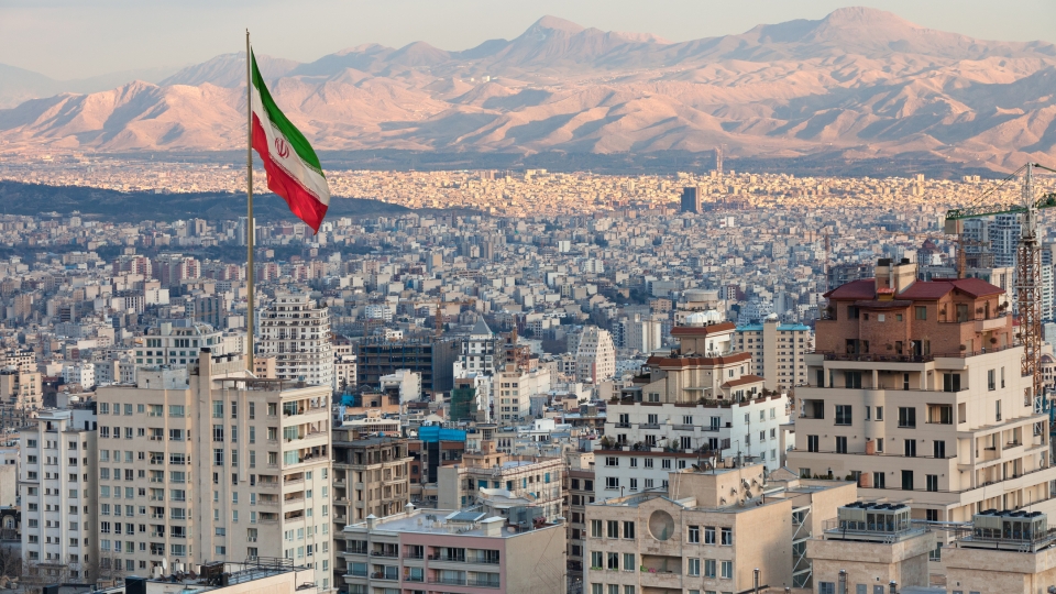 Waving Iran flag above skyline of Tehran at sunset.