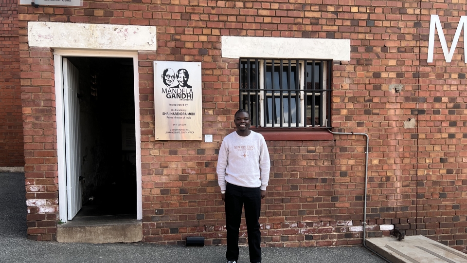 A man in a light grey sweatshirt and black pants stands in front of a red brick building at Constitution Hill in Johannesburg, next to an open doorway labeled "Isolation Cells" and a plaque commemorating the "Mandela Gandhi" exhibition.