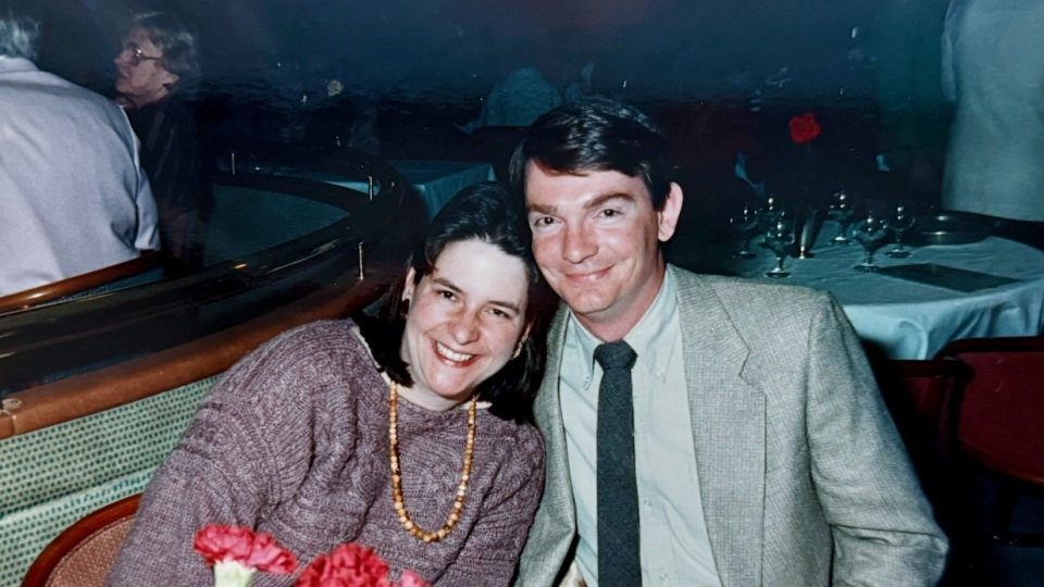 A smiling man in a light grey blazer and tie sits beside a woman in a purple knit sweater and beaded necklace at a restaurant table with red carnations.