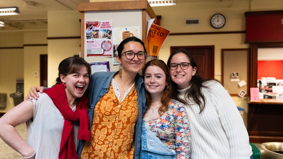 Four smiling young women pose closely together in a hallway decorated with community event posters.