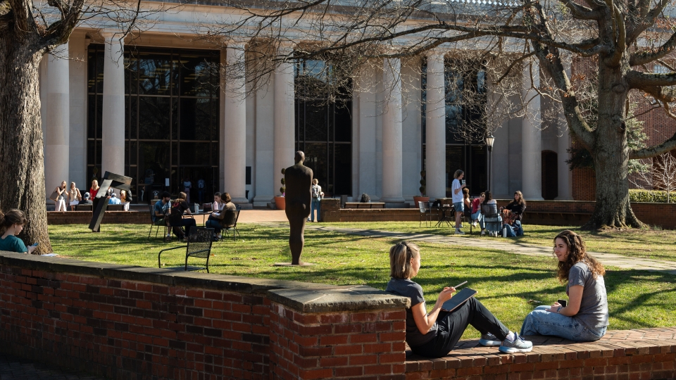 Students sit on a brick wall and at tables in a sunny campus courtyard featuring a large neoclassical building with white columns and an outdoor sculpture.