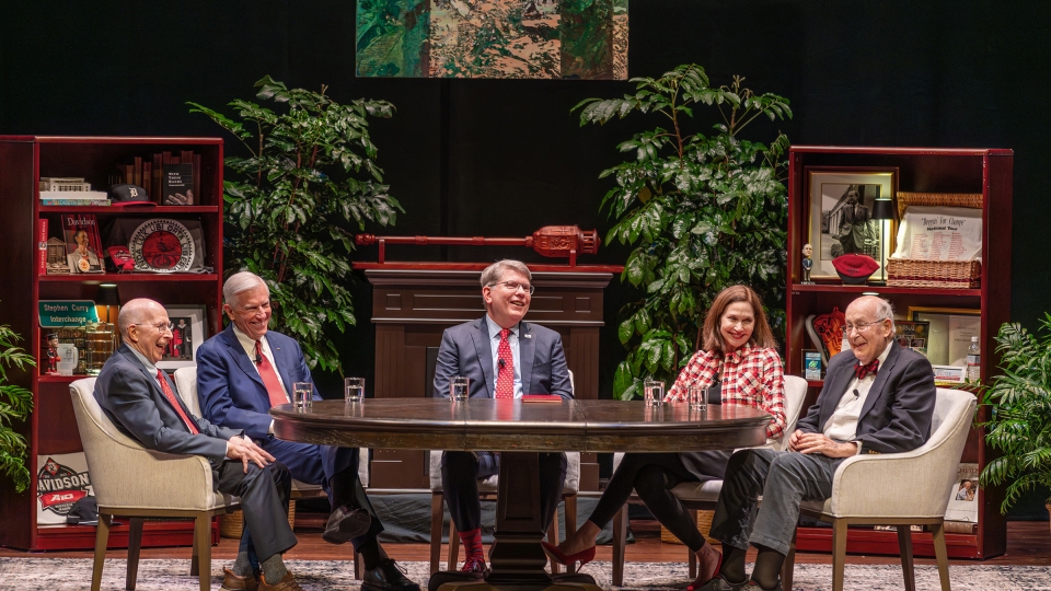 Five people sit around a large oval table on a stage set designed to look like a study, featuring bookshelves filled with memorabilia, potted plants, and an abstract painting hanging in the center.