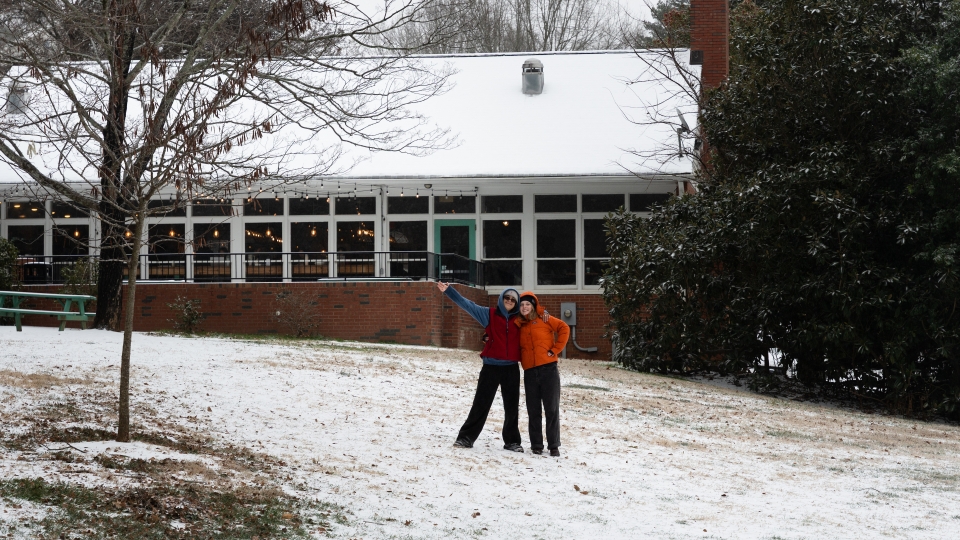 Two people wearing colorful winter jackets stand with arms around each other on a snow-dusted grassy slope in front of a brick building with a snow-covered roof and a large evergreen tree.