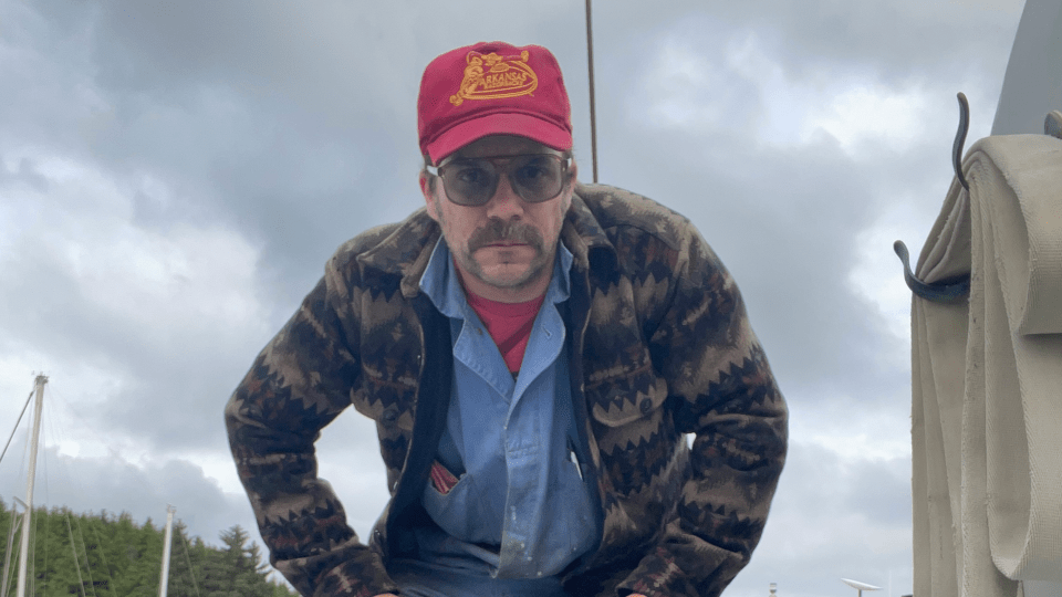 A man with a mustache, sunglasses, and a red Arkansas Razorbacks hat stands outdoors under a cloudy sky wearing a patterned wool jacket over a blue button-down shirt.