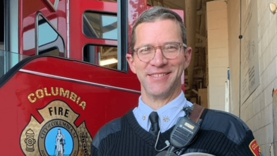 A smiling man in a black sweater and tie stands in front of a red fire truck featuring the "Columbia Fire" logo.