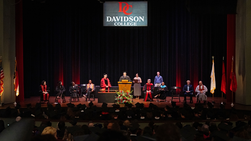 A wide view of a formal convocation ceremony on a stage with a "Davidson College" logo backdrop, featuring several faculty members in academic regalia seated around a student speaking at a podium
