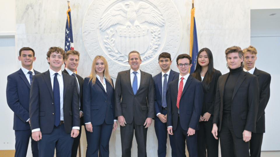 A group of ten professionally dressed students and a central official pose for a formal photo in front of a large, white architectural seal of the Federal Reserve Board of Governors.