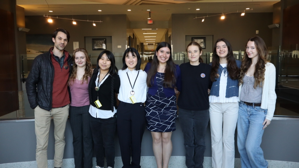 Seven young people stand side-by-side smiling in a brightly lit modern hallway with a decorative artistic ceiling.
