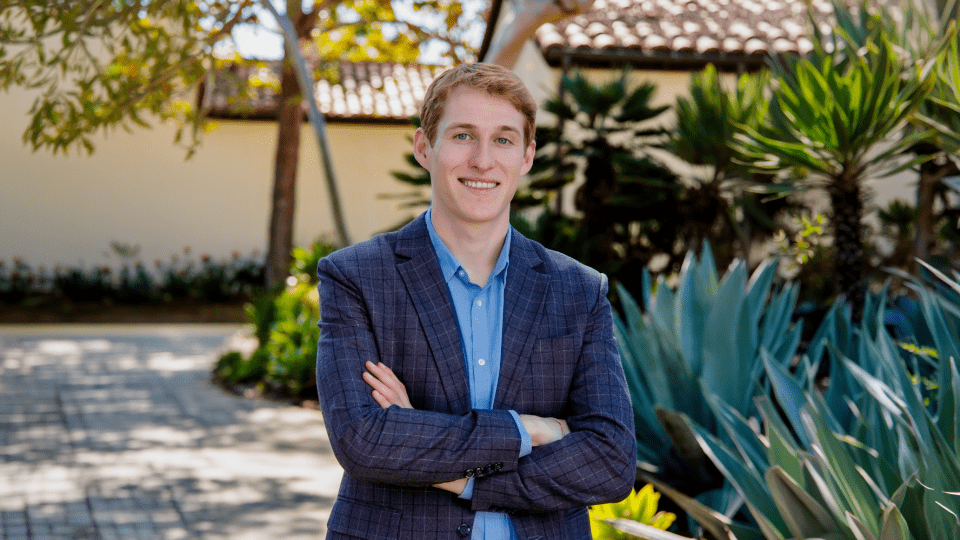 A young white man wearing a blue blazer in a tropical setting, arms crossed