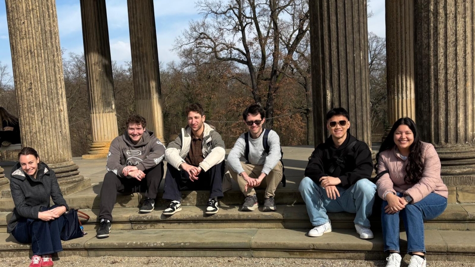 Six individuals sit smiling on the stone steps of a classical pavilion featuring large, fluted columns that frame a view of leafless trees under a clear blue sky.