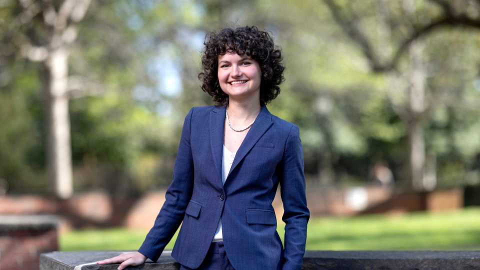 A woman with short curly hair smiles while wearing a navy blue suit and standing outdoors in front of a brick wall and blurred greenery.