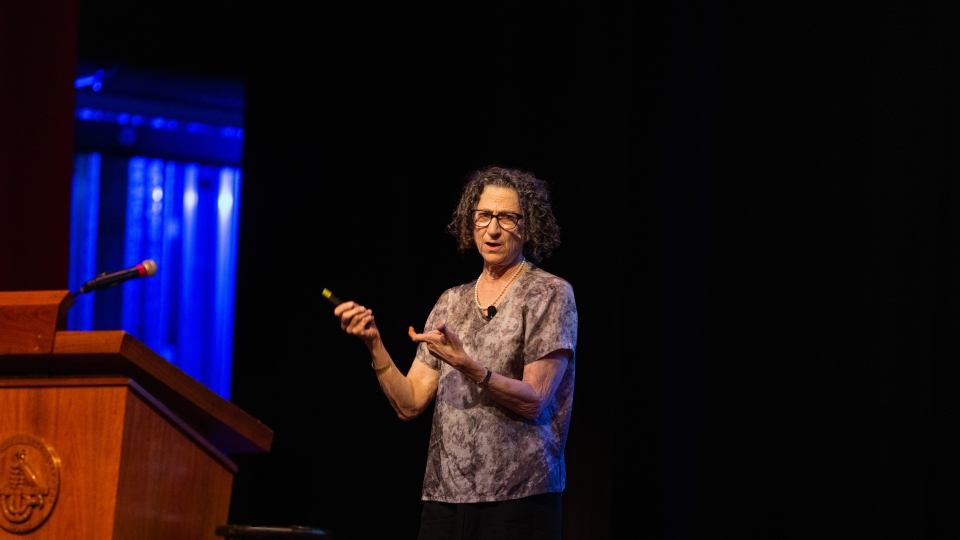 Dr. Cynthia Dwork gestures with a handheld clicker while delivering a lecture on a dimly lit stage next to a wooden podium.