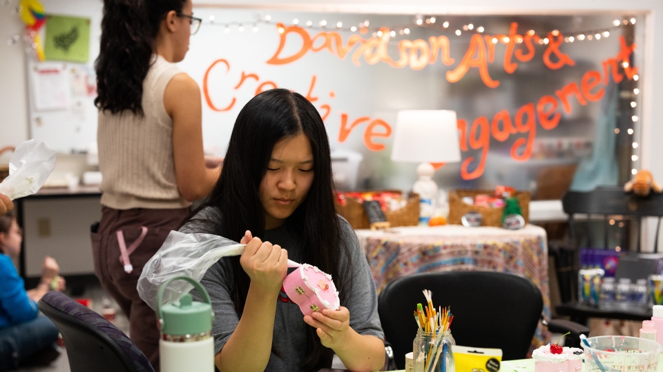 A student focused on her work uses a piping bag to apply white decorative frosting to a small heart-shaped pink jewelry box in a creative studio labeled "Davidson Arts & Creative Engagement."