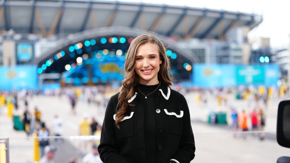 Courtney smiles at a desk with a stadium and large outdoor event stage blurred in the background.