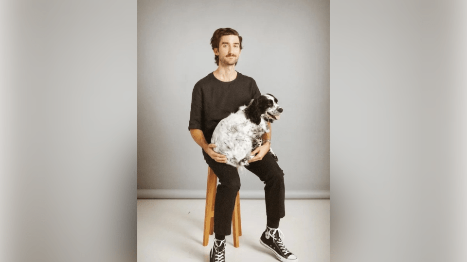 A man with a mustache sits on a wooden stool against a gray backdrop, wearing a black t-shirt, black pants, and black high-top sneakers while cradling a fluffy, black-and-white speckled dog on his lap.