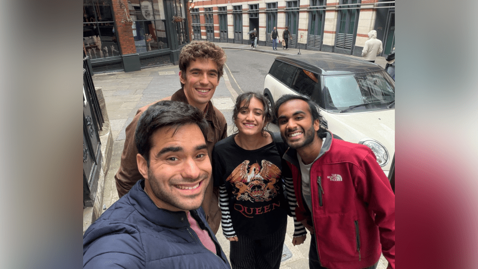 Four smiling friends take a high-angle selfie on a curved London street in front of a white compact car and a building labeled "Pub & Eating House."