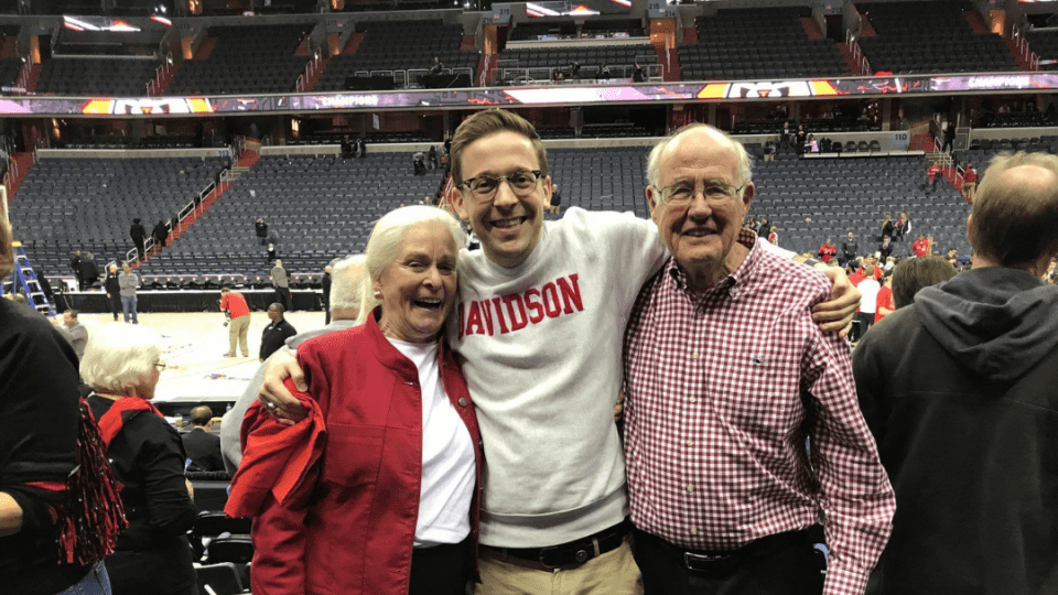 A smiling young man in a white Davidson sweatshirt stands with his arms around an older woman in a red jacket and an older man in a checkered shirt in a basketball arena.