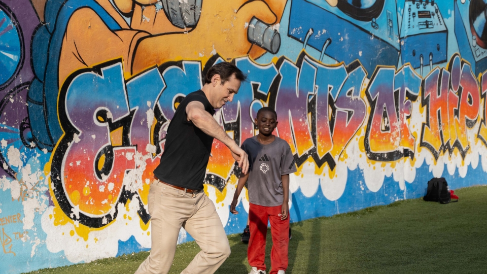 An adult man plays soccer with a young boy in front of a colorful graffiti mural