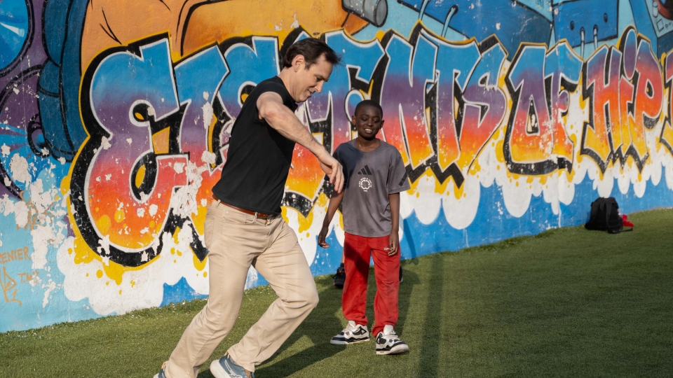 An adult man plays soccer with a young boy in front of a colorful graffiti mural