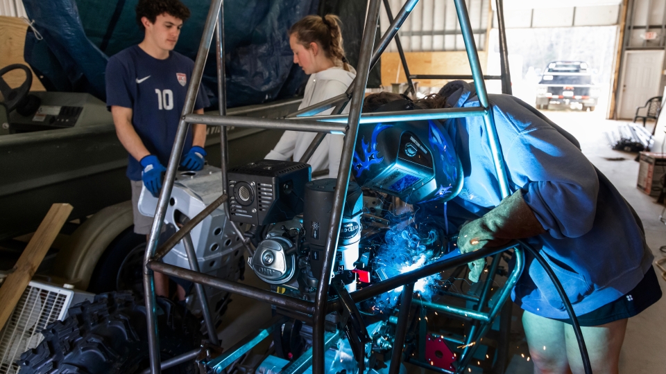 A student wearing a blue welding helmet and protective gloves welds a component onto the metal chassis of a custom off-road vehicle while two peers observe.