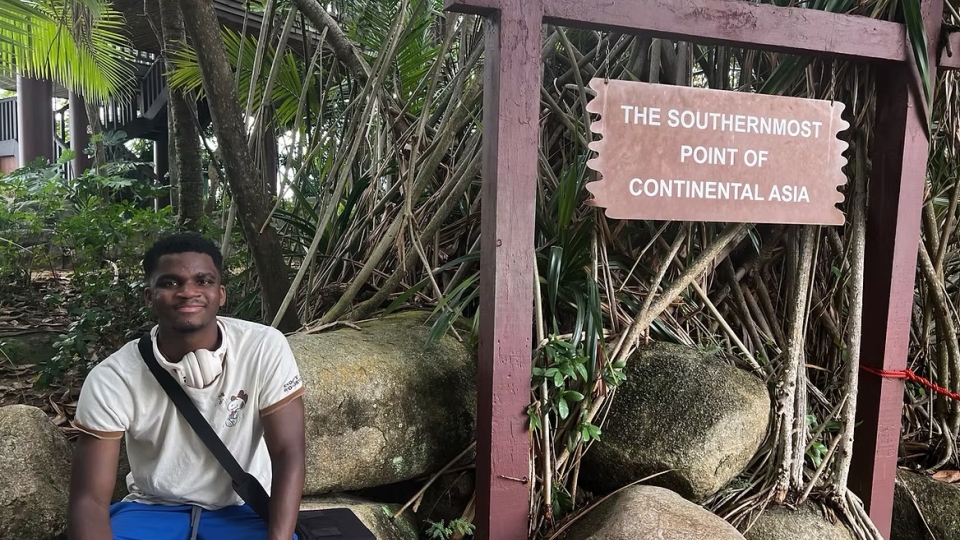 A young man sits on large rocks next to a wooden signpost reading "The Southernmost Point of Continental Asia," surrounded by tropical foliage.