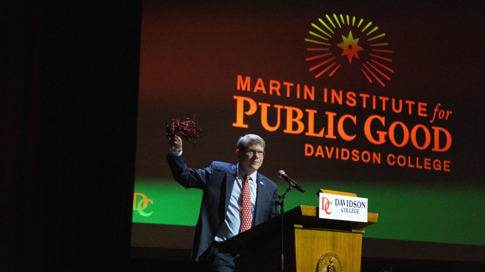 A man in a suit stands at a Davidson College podium on a dimly lit stage, raising a pom-pom in one hand beneath a large projection screen that reads "Martin Institute for Public Good."