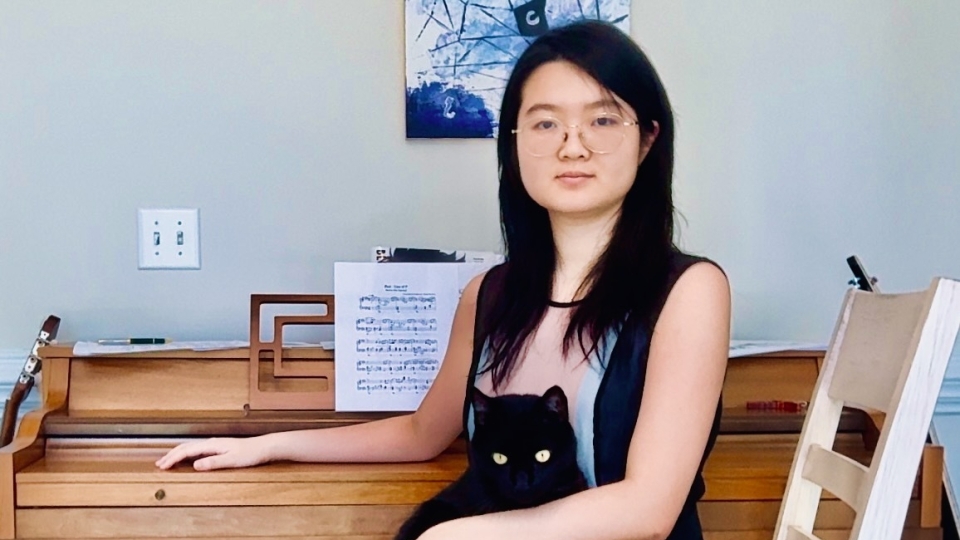 A female student with long black hair and glasses sits in front of a piano, holding a black cat