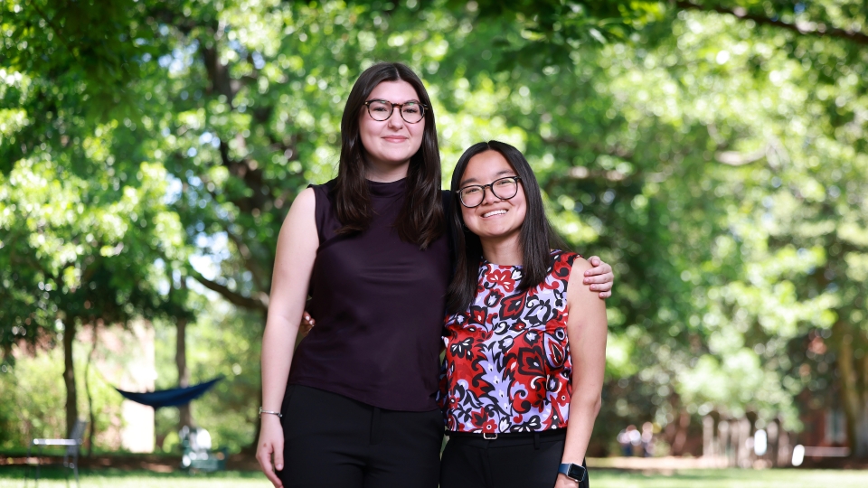 Two smiling students wearing glasses stand side-by-side with arms around each other on a sunny, green campus lawn surrounded by trees.