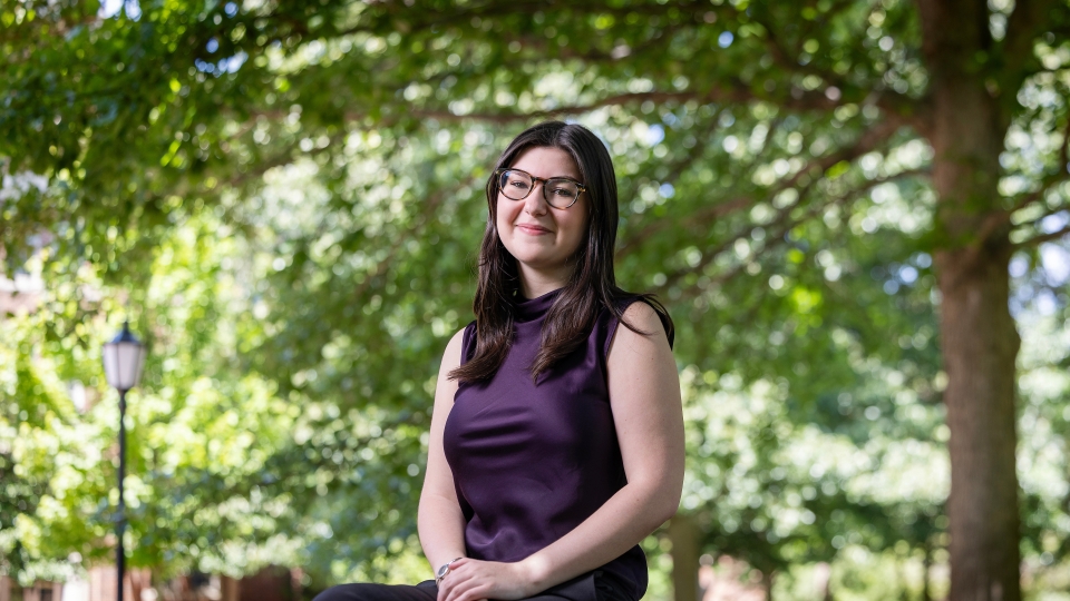 A smiling student wearing glasses and a dark purple sleeveless top sits outside on a stone ledge beneath leafy green trees.