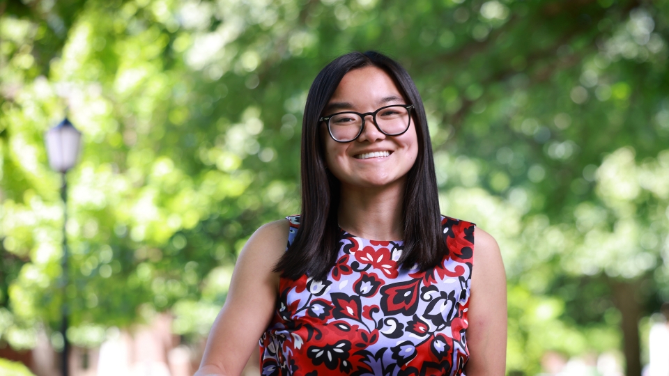 A smiling student with glasses and straight, dark hair poses outside wearing a sleeveless floral patterned blouse.
