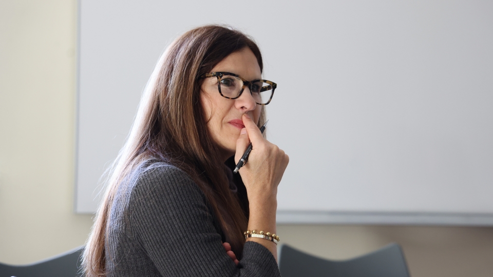 A woman with long brown hair and tortoise-shell glasses sits in a classroom, looking thoughtfully to the side while holding a pen near her lips, with a blank whiteboard in the background.