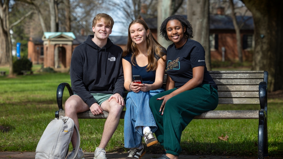 Three students sitting together on a wooden park bench, smiling at the camera.