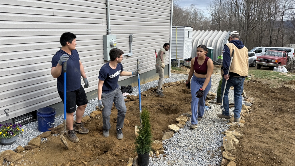 A group of volunteers works together on an outdoor landscaping project next to a house, digging trenches and laying gravel and stone borders.