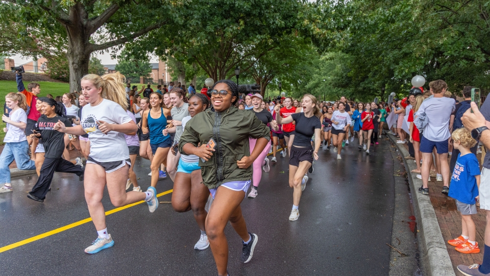 A large group of smiling young runners race down a tree-lined paved road while spectators cheer along the sidewalk.