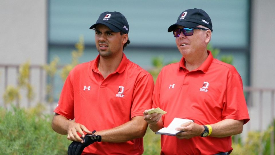 A young golfer adjusting a black glove next to a coach reviewing notes on a small yardage book. Both are wearing matching red Under Armour polo shirts and black caps branded with a white "D" and "GOLF" logo.