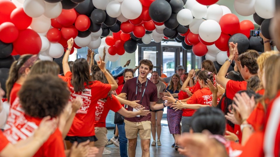 A young man walking down a hallway filled with red, white, and black balloons while crowd members in matching red shirts cheer and high-five him.