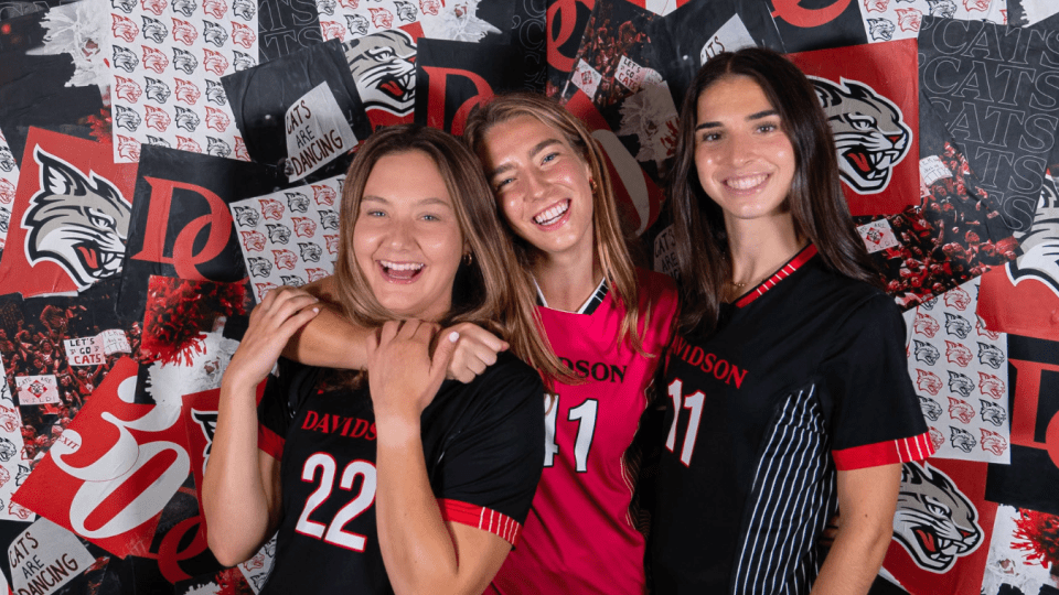 Three smiling female soccer players in Davidson College jerseys pose together in front of a red and black branded backdrop.