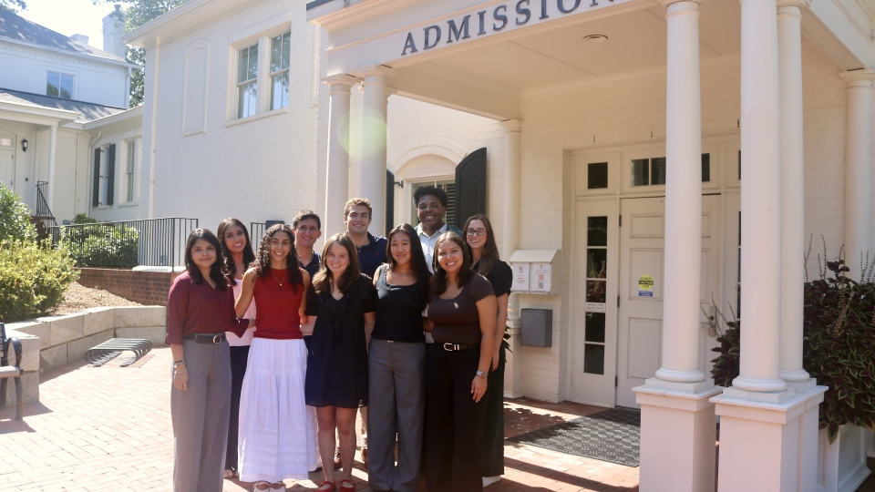 A diverse group of nine students smiling together for a group photo under the columned portico of a white "Admission" building.