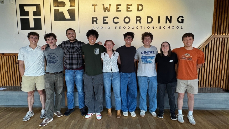 A group of eight young adults stands in a line with their arms around each other's shoulders, posing for a photo inside a modern room in front of a wall with a logo that reads "Tweed Recording Audio Production School."