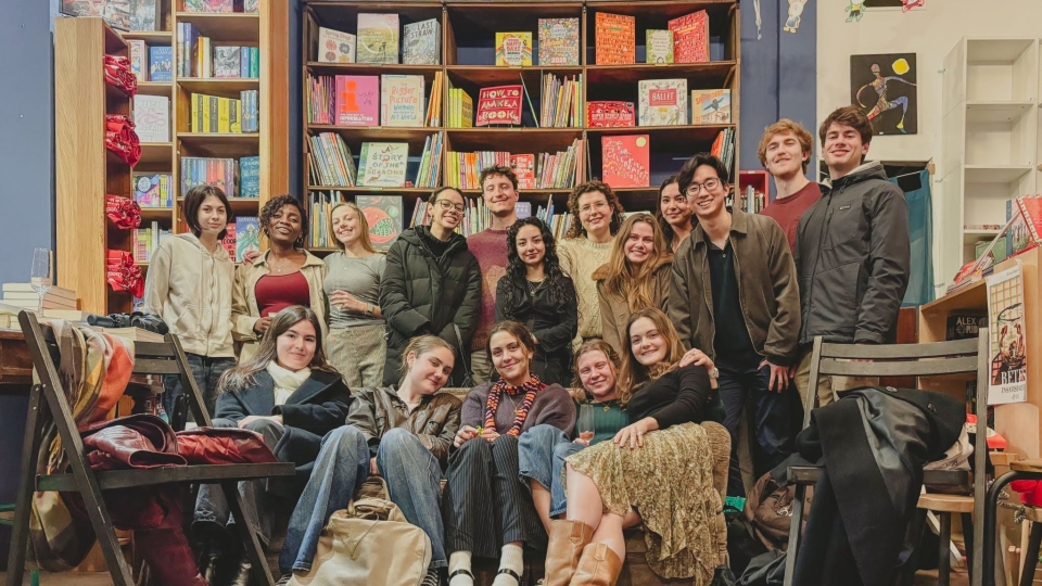 A diverse group of smiling young adults poses together inside a cozy bookstore with tall shelves filled with children's books and illustrations in the background.