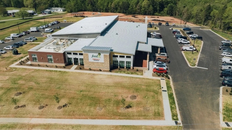 Aerial view of a large modern building with a metal roof featuring the FeedNC logo, surrounded by parking lots with parked cars, paved walkways, and open grassy areas.