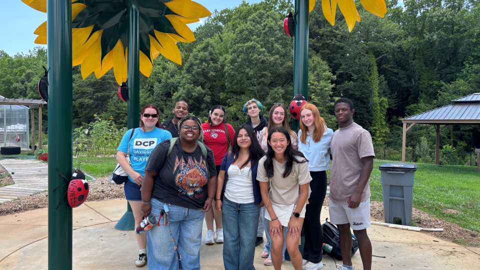 A group of young adults smiling together outdoors under giant artificial sunflower sculptures decorated with large ladybugs.