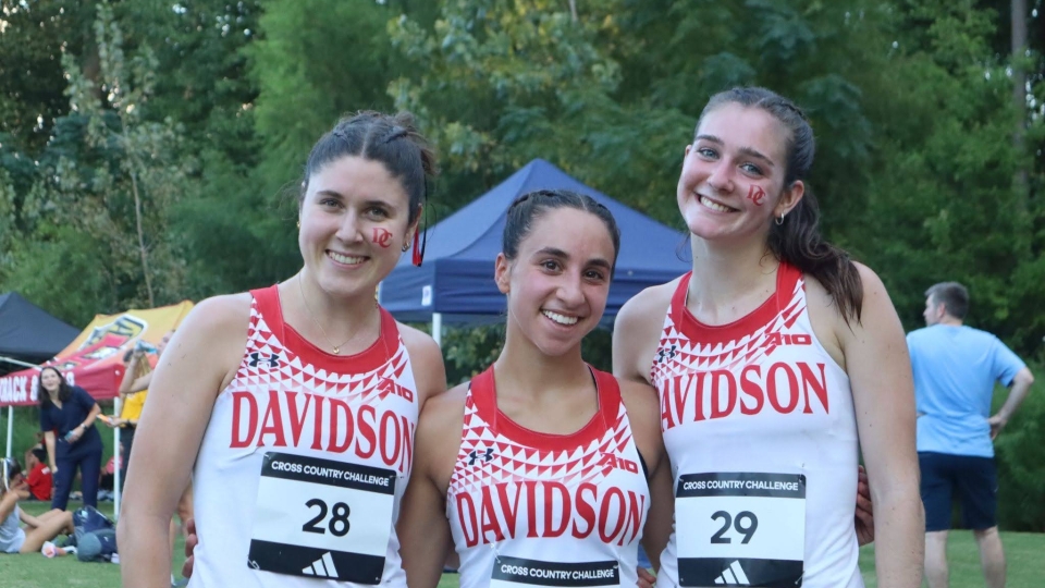 Three smiling female cross-country runners wearing red and white Davidson College uniforms and race bibs stand together outdoors.