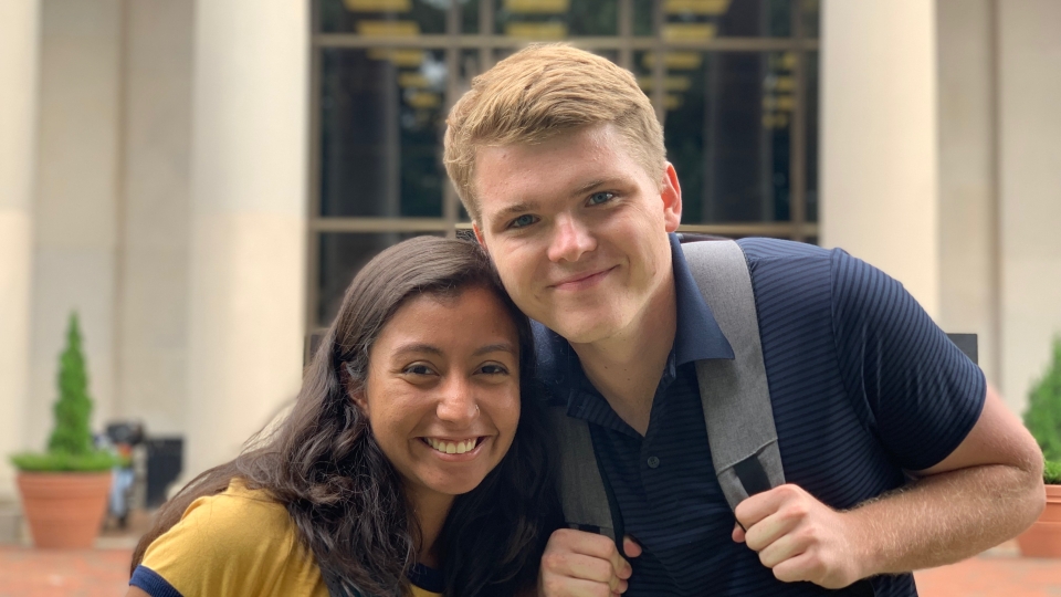 Zach and Yara smile together while wearing backpacks outdoors in front of a stone campus building with large glass windows.