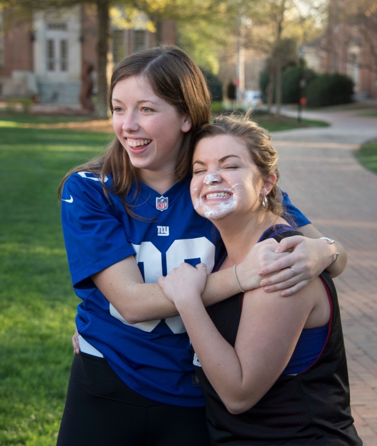 Two friends hug outside, one with pie on her face